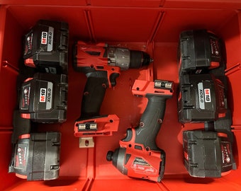 Può includere: Una cassetta degli attrezzi rossa con sei batterie nere e rosse etichettate "M18 XC6.0" e due trapani a batteria rossi e neri. Un trapano ha una funzione di martello perforatore e l'altro è un avvitatore.