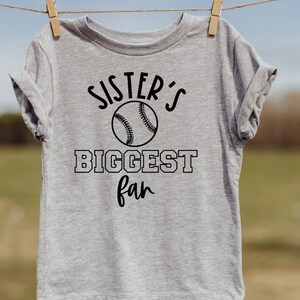 Peut inclure: T-shirt gris avec un texte noir qui dit "Sister's Biggest Fan" avec un graphique de baseball.