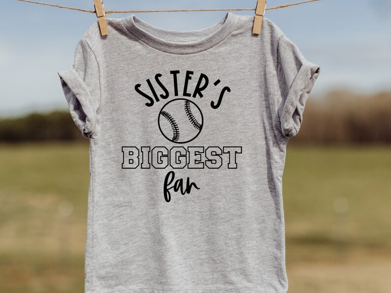 Peut inclure: T-shirt gris avec un texte noir qui dit "Sister's Biggest Fan" avec un graphique de baseball.