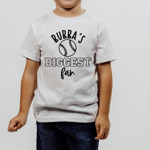 Peut inclure: T-shirt blanc avec du texte noir qui dit "Bubba's Biggest Fan" et un graphique de baseball.