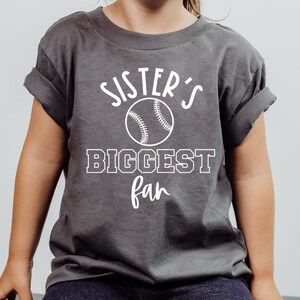K&ouml;nnte beinhalten: Ein graues T-Shirt mit wei&szlig;em Text, der "Sister's Biggest Fan" lautet, mit einem Baseball-Motiv.