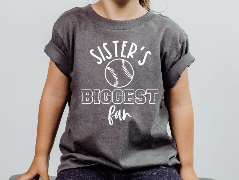 K&ouml;nnte beinhalten: Ein graues T-Shirt mit wei&szlig;em Text, der "Sister's Biggest Fan" lautet, mit einem Baseball-Motiv.