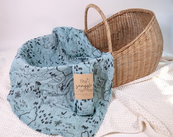May include: A light blue muslin swaddle blanket with a map design and the text "The snuggle is real" rolled up next to a wicker basket. The blanket is inside a wicker basket on a white knitted blanket.
