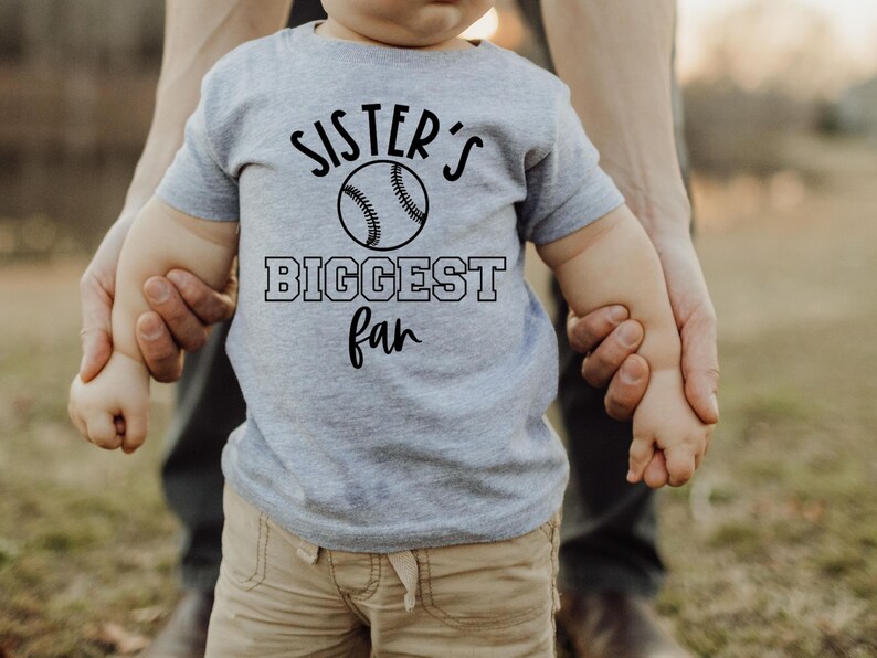 K&ouml;nnte beinhalten: Ein Kleinkind tr&auml;gt ein graues T-Shirt mit dem Text "Sister's Biggest Fan" und einer Baseballgrafik. Das Kleinkind h&auml;lt die Hand eines Erwachsenen.