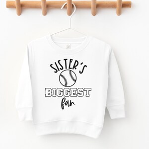Peut inclure: Sweat-shirt blanc &agrave; manches longues avec un texte noir qui dit "Sister's Biggest Fan" et un graphique de baseball noir.