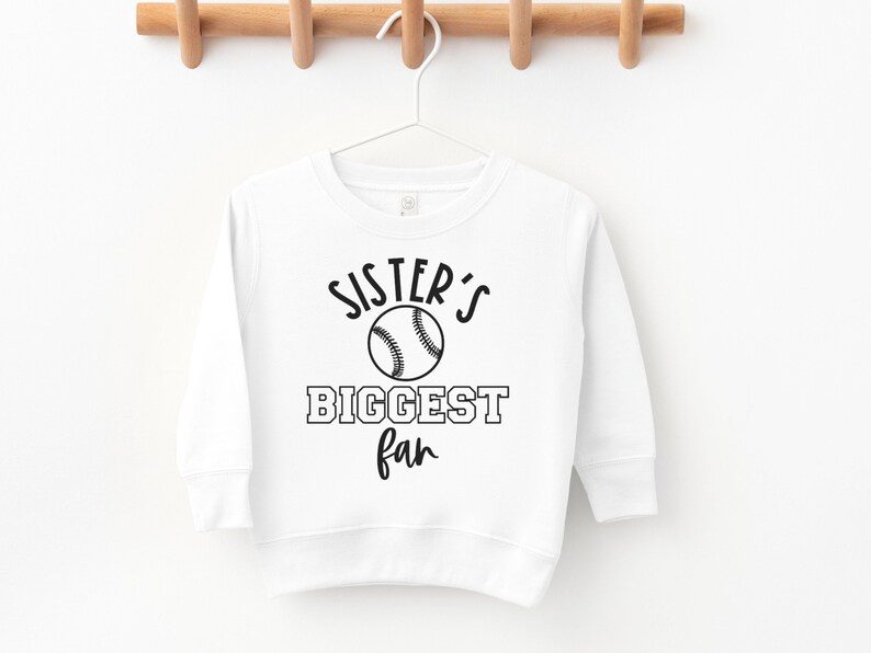 Peut inclure: Sweat-shirt blanc &agrave; manches longues avec un texte noir qui dit "Sister's Biggest Fan" et un graphique de baseball noir.