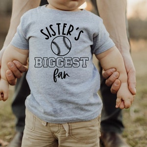 Peut inclure: Un tout-petit portant un t-shirt gris avec le texte "Sister's Biggest Fan" et un graphique de baseball. Le tout-petit tient la main d'un adulte.