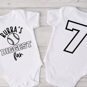 K&ouml;nnte beinhalten: Wei&szlig;er Baby-Strampler mit schwarzem Text "Bubba's Biggest Fan" und einem Baseball-Motiv. Die R&uuml;ckseite des Stramplers hat eine schwarze Nummer 7.