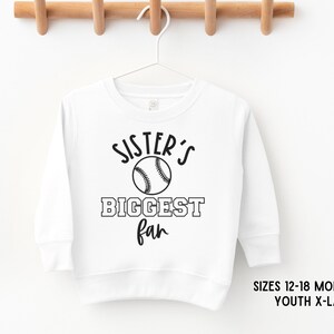 K&ouml;nnte beinhalten: Wei&szlig;er Langarm-Sweatshirt mit schwarzem Text, der "Sister's Biggest Fan" lautet, und einer Baseball-Grafik.