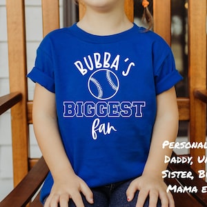 K&ouml;nnte beinhalten: Ein k&ouml;nigsblaues T-Shirt mit wei&szlig;em Text, der "Bubba's Biggest Fan" lautet, und einer wei&szlig;en Baseballgrafik. Der Text "Personalisiert: Papa, Onkel, Schwester, Bubba, Mama usw..." ist ebenfalls auf dem T-Shirt zu sehen.