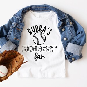 K&ouml;nnte beinhalten: Wei&szlig;es T-Shirt mit schwarzem Text, der "Bubba's Biggest Fan" mit einem Baseball-Motiv lautet. Ein brauner Leder-Baseballhandschuh mit einem wei&szlig;en Baseball befindet sich in der linken unteren Ecke des Bildes. Eine blaue Jeansjacke ist auf dem T-Shirt gefaltet.