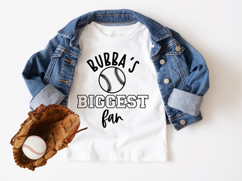 K&ouml;nnte beinhalten: Wei&szlig;es T-Shirt mit schwarzem Text, der "Bubba's Biggest Fan" mit einem Baseball-Motiv lautet. Ein brauner Leder-Baseballhandschuh mit einem wei&szlig;en Baseball befindet sich in der linken unteren Ecke des Bildes. Eine blaue Jeansjacke ist auf dem T-Shirt gefaltet.