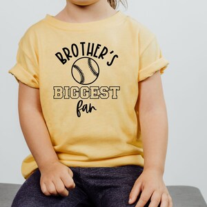 Peut inclure: Un t-shirt jaune avec du texte noir qui dit "Brother's Biggest Fan" et un graphique de baseball.