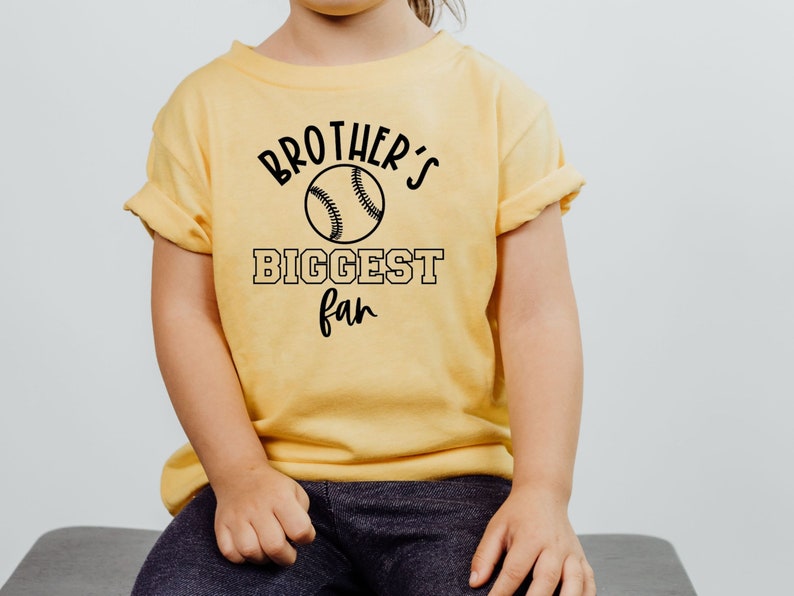Peut inclure: Un t-shirt jaune avec du texte noir qui dit "Brother's Biggest Fan" et un graphique de baseball.