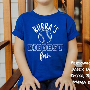 Peut inclure: Un t-shirt bleu royal avec du texte blanc qui dit "Bubba's Biggest Fan" et un graphique de baseball blanc. Le texte "Personnalis&eacute; : Papa, Oncle, S&oelig;ur, Bubba, Maman ETC..." est &eacute;galement visible sur le t-shirt.