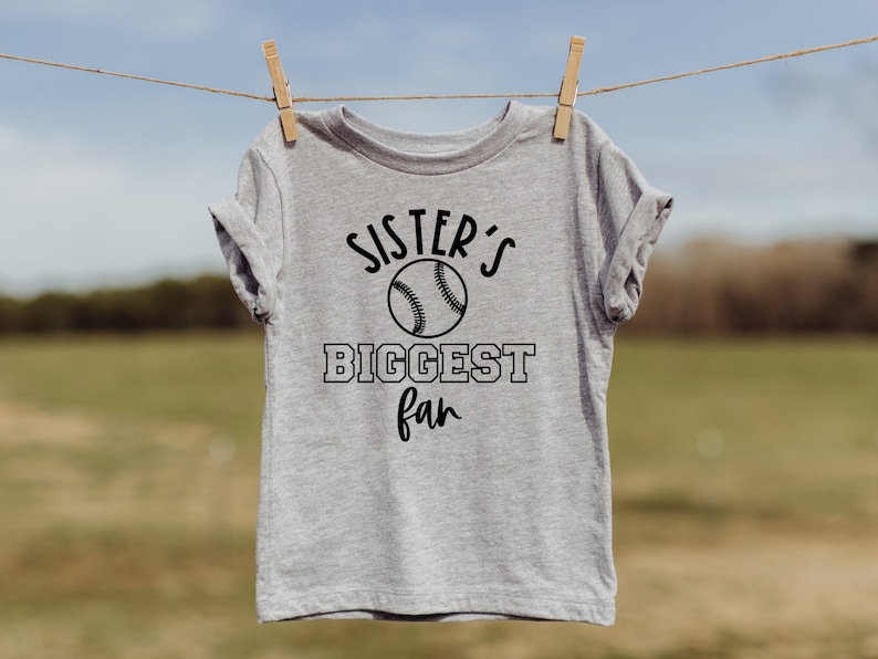 K&ouml;nnte beinhalten: Ein graues T-Shirt mit einem schwarzen Baseball-Motiv und dem Text "Sister's Biggest Fan".