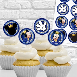 Puede incluir: Toppers para cupcakes azules y dorados con Mickey Mouse vestido de príncipe. Los toppers están encima de cupcakes con glaseado blanco. La imagen también incluye una hoja de toppers para cupcakes imprimibles con varios diseños.