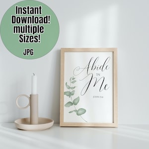 Op de afbeelding: Een ingelijst print met een witte achtergrond en een zwart kalligrafie citaat dat "Abide in Me" luidt, met de bijbelverwijzing "Johannes 15:4" eronder. De print bevat een aquarel illustratie van groene eucalyptusbladeren in de hoek.