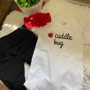 Puede incluir: Un body blanco con una mariquita negra y el texto "cuddle bug" en la parte delantera. El body se combina con pantalones negros con volantes y una diadema de flores rojas con un adorno de pedrería.