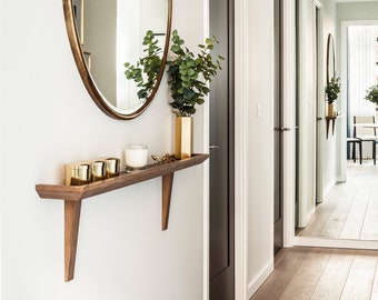 Könnte beinhalten: Ein Flur mit weißen Wänden und dunklem Holzboden. Ein Holzregal mit goldenen Kerzen und einer Vase mit Grünpflanzen ist an der Wand montiert. Ein großer ovaler Spiegel mit goldenem Rahmen hängt über dem Regal. Zwei geschlossene Türen mit dunklen Holzrahmen sind auf der linken Seite des Bildes zu sehen. Ein Spiegel auf der rechten Seite des Bildes spiegelt den Flur wider.