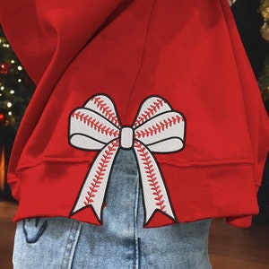 May include: Red sweatshirt with a white baseball-stitched bow on the back. The bow is positioned to look like it is tied around the wearer's waist.