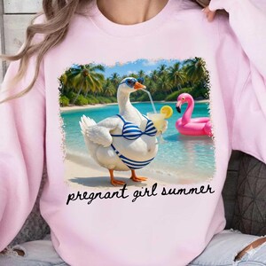 Pu&ograve; includere: Una felpa rosa chiaro con un'immagine di un'oca in bikini a righe blu e bianche e occhiali da sole, che tiene in mano un cocktail su una spiaggia. Un galleggiante a forma di fenicottero rosa sullo sfondo. Il testo "pregnant girl summer" &egrave; sotto.