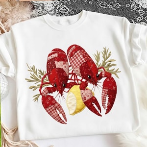May include: White crew neck sweatshirt featuring a red crawfish design with a lemon slice and sprigs of greenery. The crawfish are detailed with red and white patterned fabric. A fun and unique design.