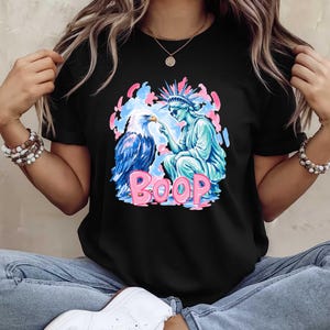 May include: Black t-shirt with a graphic design featuring a blue and white eagle, a turquoise Statue of Liberty, and the pink word "BOOP." The design is set against pink and blue clouds.