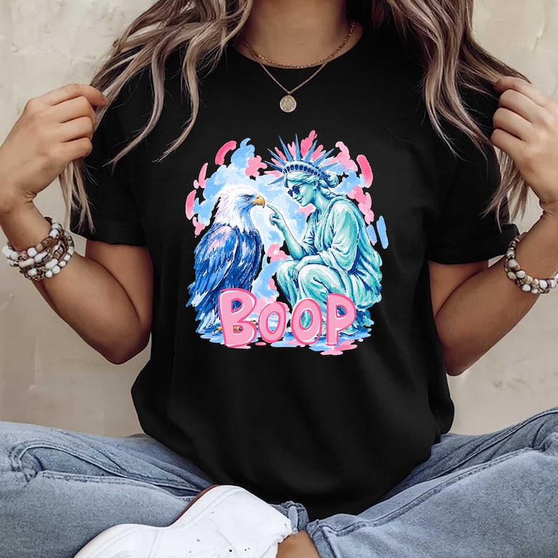 May include: Black t-shirt with a graphic design featuring a blue and white eagle, a turquoise Statue of Liberty, and the pink word "BOOP." The design is set against pink and blue clouds.