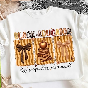 Puede incluir: Camiseta blanca con la frase "BLACK EDUCATOR" en letras marrones y doradas. Debajo, tres diseños rectangulares con lazos, libros y una manzana. La frase "by popular demand" está escrita en cursiva negra.