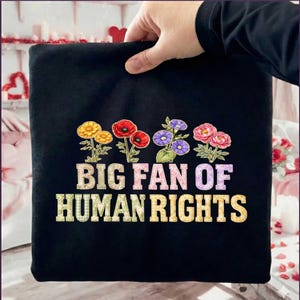 Könnte beinhalten: Schwarzes T-Shirt mit floralem Design und dem Text "BIG FAN OF HUMAN RIGHTS" in einer farbenfrohen, strukturierten Schriftart. Die Blumen umfassen gelbe, rote, violette und rosa Blüten.