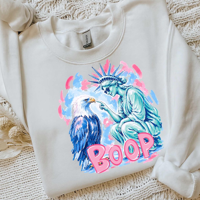 May include: White sweatshirt with a graphic of the Statue of Liberty and an eagle, with the word "BOOP" in pink. The Statue of Liberty is blue, and the eagle has blue and white feathers. Pink and blue brushstrokes surround the figures.