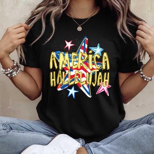 May include: Black t-shirt featuring a patriotic design with the words "AMERICA HALLELUJAH" in gold lettering. The design includes a star with red, white, and blue stripes and several smaller stars in various colors.