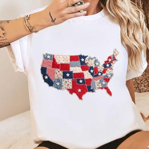 May include: White t-shirt featuring a patchwork design of the United States map. The map is composed of red, white, and blue fabric pieces with star accents. The shirt has short sleeves and a crew neck.