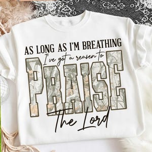 Könnte beinhalten: Weißes Sweatshirt mit dem Text "AS LONG AS I'M BREATHING I've got a reason to PRAISE The Lord." Das Wort "PRAISE" ist in großen Blockbuchstaben mit einem Camouflagemuster gehalten. Das Sweatshirt hat einen Rundhalsausschnitt und kurze Ärmel.