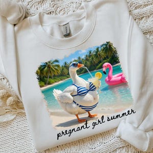Pu&ograve; includere: Felpa bianca con una grafica di un'oca con occhiali da sole e un bikini a righe blu e bianche, che tiene in mano un cocktail su una spiaggia. Un galleggiante a forma di fenicottero rosa &egrave; sullo sfondo. Il testo "pregnant girl summer" &egrave; sotto.