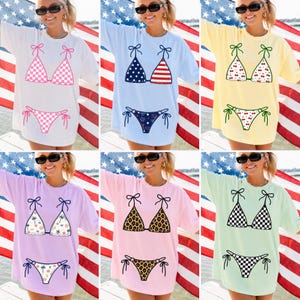 May include: A collection of oversized t-shirts featuring bikini designs. The shirts come in various colors, including white, blue, yellow, pink, and green. Bikini patterns include checkered, American flag, cherry, palm tree, leopard print, and checkered patterns.