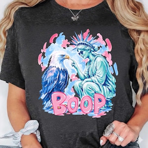 May include: Dark grey t-shirt with a graphic of an eagle and the Statue of Liberty. The eagle is blue and white, and the statue is turquoise. The word "BOOP" is in pink. The design is surrounded by pink and blue accents.