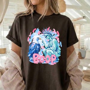 May include: Dark brown t-shirt with a graphic of an eagle and the Statue of Liberty, with the word "BOOP" in pink. The eagle is blue and white, and the Statue of Liberty is light blue. The background has pink and blue accents.