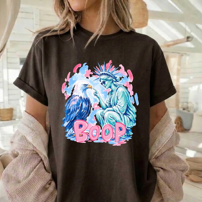 May include: Dark brown t-shirt with a graphic of an eagle and the Statue of Liberty, with the word "BOOP" in pink. The eagle is blue and white, and the Statue of Liberty is light blue. The background has pink and blue accents.
