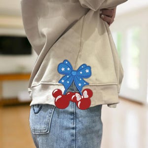 Könnte beinhalten: Ein beiger Pullover mit einer blauen Schleife mit weißen Sternen und roten Kirschen mit weißen Schleifen am unteren Saum.