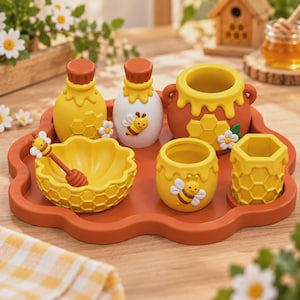 Peut inclure: Un ensemble de cuisine sur le thème du miel sur un plateau orange festonné, comprenant deux bouteilles, un grand pot à miel, un petit bol avec une cuillère à miel, une tasse jaune et un récipient hexagonal, ornés d'alvéoles et d'abeilles.