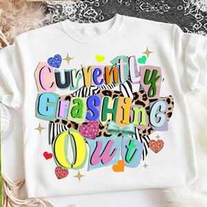 May include: White sweatshirt with the phrase "Currently Crashing Out" in colorful, layered text. The design includes hearts, stars, and animal print patterns. A fun and vibrant graphic tee for casual wear.