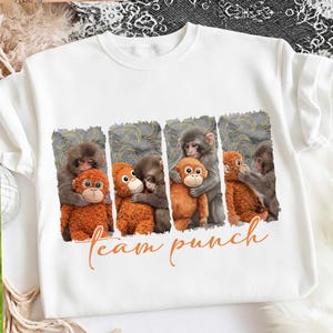 May include: White sweatshirt featuring a graphic of several monkeys hugging orange plush monkeys. The phrase "team punch" is written in orange script below the image. The sweatshirt is made of a soft material.