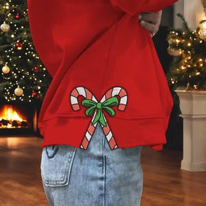 May include: A red sweatshirt with an embroidered candy cane design. The candy canes are red and white striped, tied with a green bow. The sweatshirt is paired with blue jeans, suggesting a casual, festive outfit.