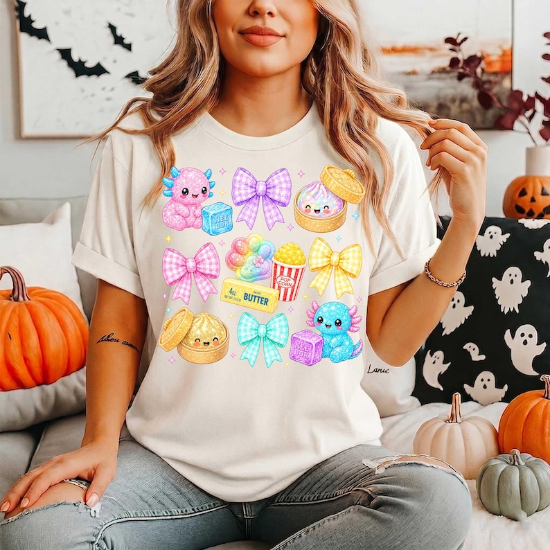 Może przedstawiać: Kremowy t-shirt z kolorowym nadrukiem graficznym przedstawiającym dwa aksolotle, trzy kokardy w kratkę, odcisk łapy, kubełek popcornu, kostkę masła oraz dwa bambusowe parowniki z uśmiechniętymi buźkami w pastelowych odcieniach r&oacute;żu, błękitu i ż&oacute;łci.