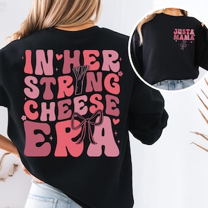 May include: Black sweatshirt featuring the text "IN HER STRING CHEESE ERA" in pink and red lettering on the back. A smaller image on the front says "JUST A MAMA" in pink.