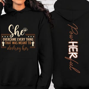 Könnte beinhalten: Schwarzer Kapuzenpullover mit dem Text "She" und "OVERCAME EVERY THING THAT WAS MEANT TO destroy her" in Gold auf der Vorderseite. Auf der Rückseite des Hoodies steht vertikal das Wort "Pow-HER ful".