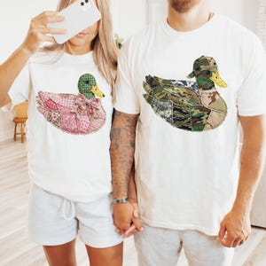 May include: White t-shirts featuring duck designs. One duck is patchwork with pink floral patterns, a green head, and a bow. The other duck has a camouflage design and a matching cap. The shirts are casual wear.