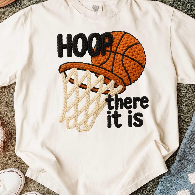 May include: Cream-colored t-shirt featuring a basketball and hoop design. The word "HOOP" is in black, above a basketball with a net. The phrase "there it is" is below the hoop in black. The basketball and hoop are in shades of brown and beige.